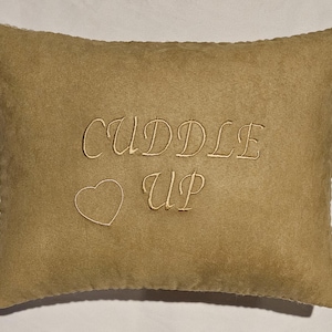 May include: A tan-colored throw pillow with the words "Cuddle Up" embroidered in a light brown color. A small heart is embroidered above the word "Up".