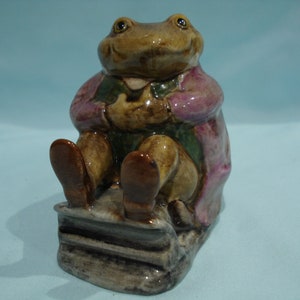 May include: A ceramic figurine of a frog wearing a pink coat and sitting on a stack of books. The frog has a green and brown body and brown eyes.