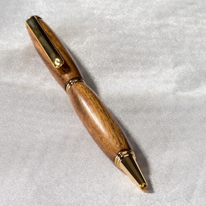 May include: A wooden pen with a gold-toned cap and tip. The pen is lying on a white fabric surface.