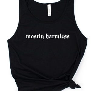 May include: A black sleeveless tank top featuring the white text mostly harmless printed in a gothic font across the chest. The garment includes a white brand label at the inner neckline and is tied in a knot at the bottom right corner.