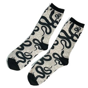 May include: A pair of crew-length socks featuring a repeating black snake pattern on a cream-coloured background. The socks have solid black cuffs, heels, and toes, with a subtle dotted texture visible throughout the light-coloured fabric sections.