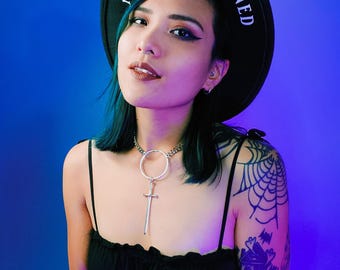 May include: A black hat with the text "MORBIDLY INCLINED" in white lettering. The person is wearing a black top and a silver sword pendant necklace. A tattoo of a spiderweb and a haunted house is visible on the shoulder.