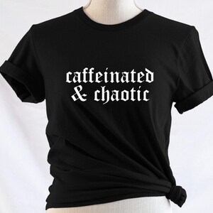 Puede incluir: Una camiseta negra de manga corta con el texto blanco "caffeinated & chaotic" impreso en una fuente de estilo gótico en el pecho. La camiseta tiene cuello redondo, mangas remangadas y el dobladillo inferior anudado a un lado.