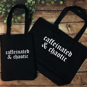 Puede incluir: Dos bolsas de tela negras con asas negras a juego, cada una con el texto blanco "caffeinated & chaotic" impreso en una fuente de estilo gótico. Una bolsa está orientada verticalmente, mientras que la otra está colocada horizontalmente.