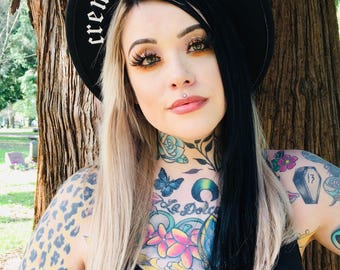 May include: A black hat with the words "cremated equal" written in white on the brim. The person wearing the hat has black and blonde hair and colorful tattoos on their neck and chest.
