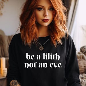 May include: Black sweatshirt with white text that reads "be a lilith not an eve". A woman with long, wavy red hair is wearing the sweatshirt.