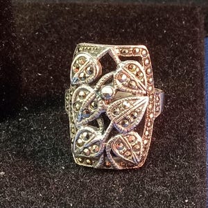 May include: Silver-toned ring with a rectangular face featuring a leaf design. The leaves are detailed with small, round, sparkling stones. The ring has a vintage or antique aesthetic, suitable for jewelry collections.