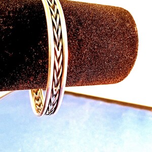 May include: A silver-colored bangle bracelet with a braided design. The bracelet is displayed on a dark brown, velvety surface. The braided pattern is visible on the outer edge of the bracelet, creating a textured appearance. The background is a light blue and white.