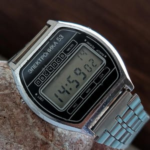 May include: A vintage digital wristwatch with a silver-tone case and a black face displaying the time. The watch has a silver band and is resting on a textured, light brown rock. The watch face has the text "ЭЛЕКТРОНИКА 53".