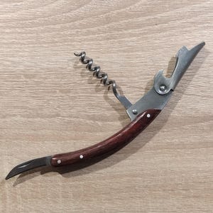 May include: A waiter's corkscrew featuring a dark brown wooden handle with two silver rivets, a stainless steel spiral worm, a serrated foil cutter blade, and a bottle opener lever mechanism. The tool is shown in an open, partially extended position.