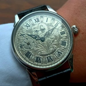 May include: Silver wristwatch with a black leather strap. The watch face displays Roman numerals and detailed engraving. A small subdial is present. The watch is worn on a wrist.