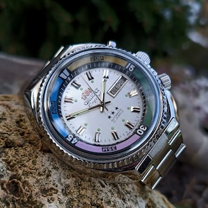 Può includere: Un orologio automatico Orient Crystal argentato con quadrante bianco, lancette nere e lunetta girevole con indicazioni per 20M, 30M, 40M e 45. L'orologio ha una finestrella per la data e una finestrella per il giorno della settimana. L'orologio è su un braccialetto in metallo argentato.