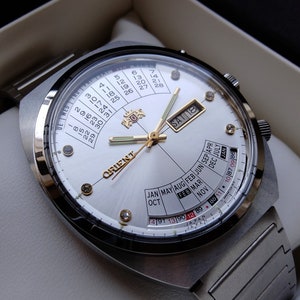 May include: A silver Orient watch with a white face and a calendar display. The watch has a silver band and a date window at the 3 o'clock position. The watch face also has a day of the week display and a month display.