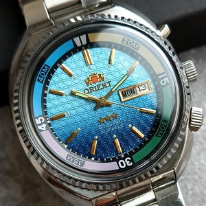 May include: A stainless steel Orient watch with a blue and teal honeycomb patterned dial, gold-tone hands and indices, and a day-date window. The bezel features a multi-colored scale with markings including 40M, 35M, 30M, 20M, and 15.