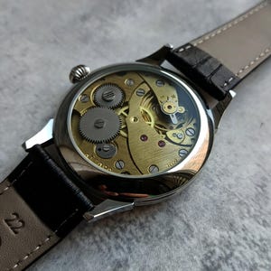 Puede incluir: Primer plano de un reloj de pulsera mec&aacute;nico con parte trasera transparente, que muestra los intrincados engranajes y mecanismos dorados. El reloj tiene una caja plateada y una correa de cuero negro. El reloj est&aacute; sobre una superficie gris.