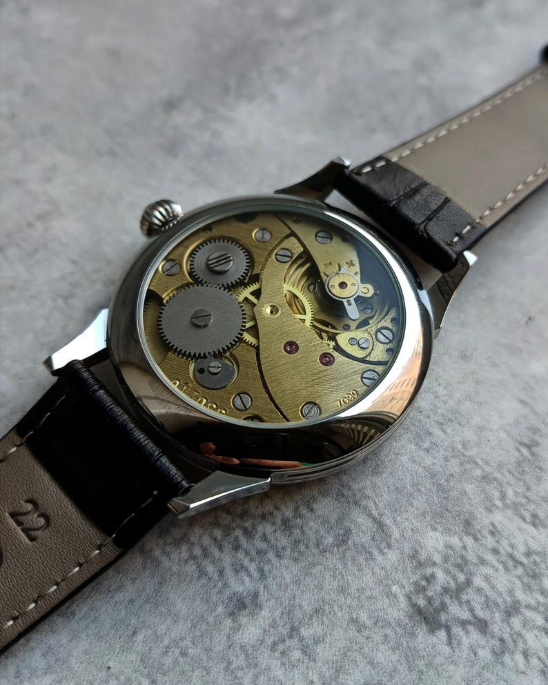 Puede incluir: Primer plano de un reloj de pulsera mec&aacute;nico con parte trasera transparente, que muestra los intrincados engranajes y mecanismos dorados. El reloj tiene una caja plateada y una correa de cuero negro. El reloj est&aacute; sobre una superficie gris.