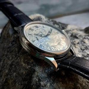 Puede incluir: Primer plano de un reloj de pulsera plateado con una esfera detallada y ornamentada con n&uacute;meros romanos. El reloj tiene una correa de cuero negro. El reloj est&aacute; sobre una superficie de piedra gris.
