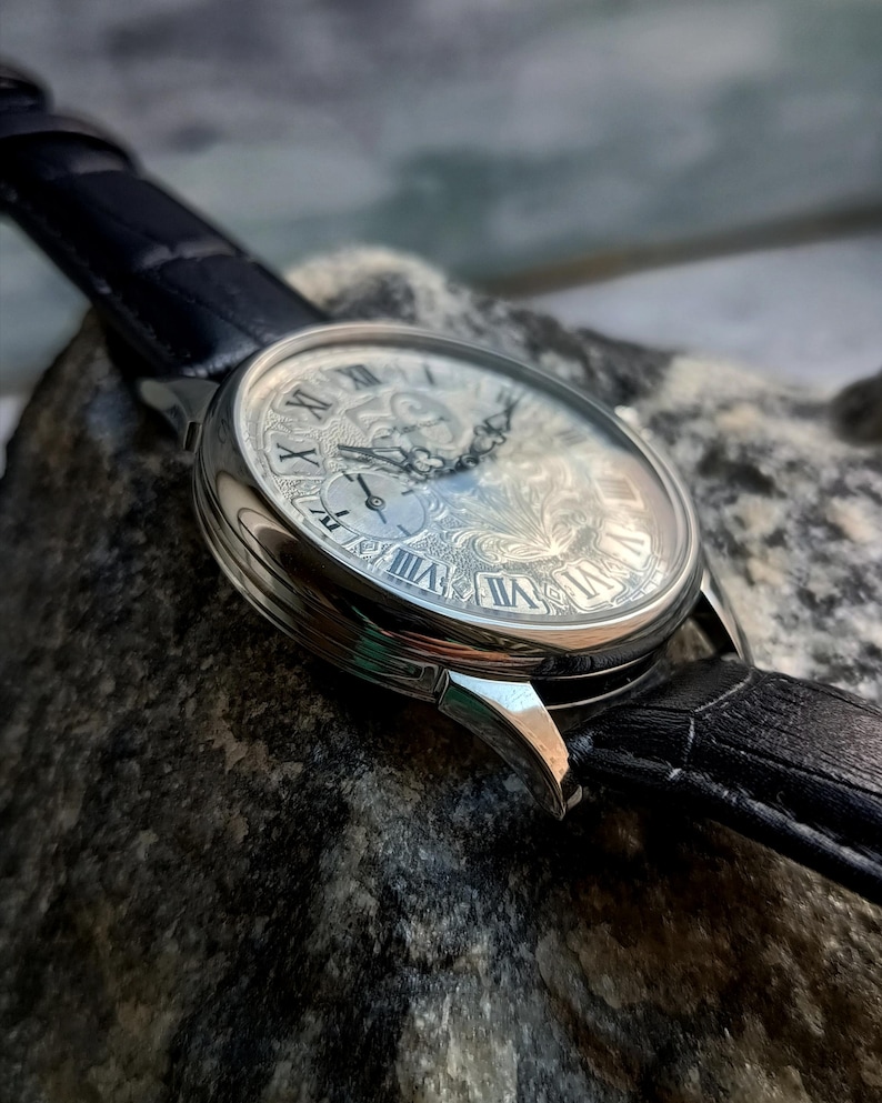 Puede incluir: Primer plano de un reloj de pulsera plateado con una esfera detallada y ornamentada con n&uacute;meros romanos. El reloj tiene una correa de cuero negro. El reloj est&aacute; sobre una superficie de piedra gris.