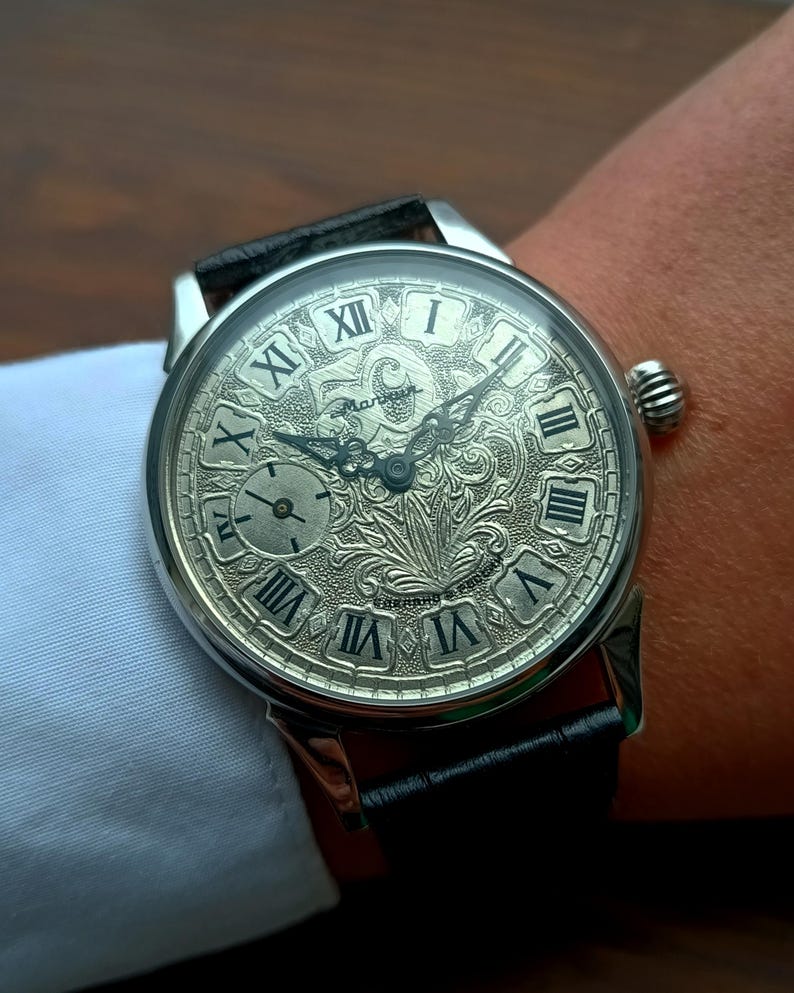 May include: Silver wristwatch with a black leather strap. The watch face displays Roman numerals and detailed engraving. A small subdial is present. The watch is worn on a wrist.
