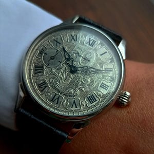 Puede incluir: Reloj de pulsera plateado con una esfera grabada con detalles, que muestra n&uacute;meros romanos y dise&ntilde;os ornamentales. El reloj tiene una correa de cuero negro y una peque&ntilde;a corona texturizada. El reloj est&aacute; en una mu&ntilde;eca, con una manga de camisa blanca en el fondo.
