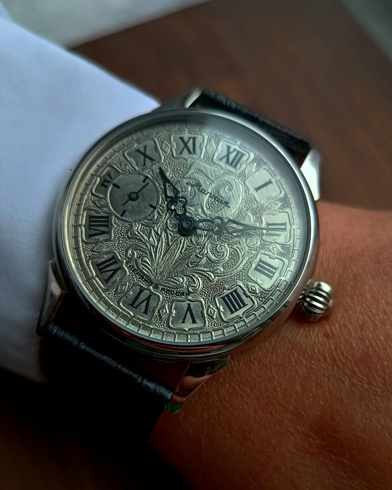 Puede incluir: Reloj de pulsera plateado con una esfera grabada con detalles, que muestra n&uacute;meros romanos y dise&ntilde;os ornamentales. El reloj tiene una correa de cuero negro y una peque&ntilde;a corona texturizada. El reloj est&aacute; en una mu&ntilde;eca, con una manga de camisa blanca en el fondo.