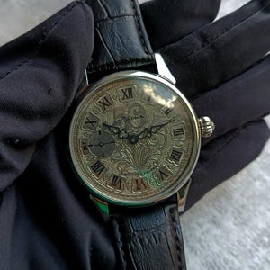 Puede incluir: Un reloj de pulsera plateado con un dise&ntilde;o floral detallado en la esfera y n&uacute;meros romanos. El reloj tiene una peque&ntilde;a subesfera y una correa de cuero negro. El reloj est&aacute; sujeto por una mano enguantada.