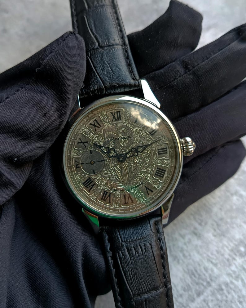 Puede incluir: Un reloj de pulsera plateado con un dise&ntilde;o floral detallado en la esfera y n&uacute;meros romanos. El reloj tiene una peque&ntilde;a subesfera y una correa de cuero negro. El reloj est&aacute; sujeto por una mano enguantada.