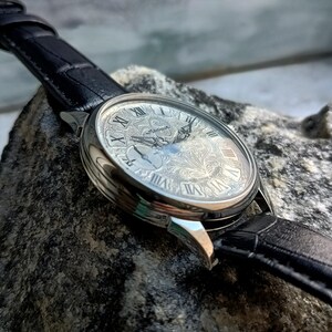 Puede incluir: Un reloj de pulsera plateado con una correa de cuero negro. La esfera del reloj presenta n&uacute;meros romanos y un grabado floral intrincado. El reloj se encuentra sobre un fondo texturizado, mostrando su dise&ntilde;o detallado.