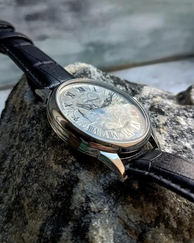 Puede incluir: Un reloj de pulsera plateado con una correa de cuero negro. La esfera del reloj presenta n&uacute;meros romanos y un grabado floral intrincado. El reloj se encuentra sobre un fondo texturizado, mostrando su dise&ntilde;o detallado.