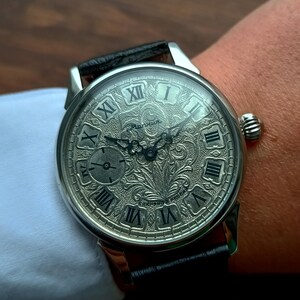 Puede incluir: Reloj de pulsera plateado con una correa de cuero negro. La esfera del reloj presenta n&uacute;meros romanos y un dise&ntilde;o grabado detallado. El reloj est&aacute; en una mu&ntilde;eca, con una manga de camisa blanca en el fondo.