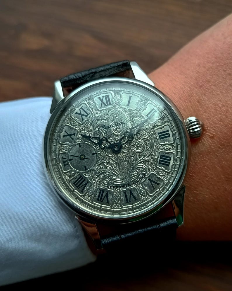 Puede incluir: Reloj de pulsera plateado con una correa de cuero negro. La esfera del reloj presenta n&uacute;meros romanos y un dise&ntilde;o grabado detallado. El reloj est&aacute; en una mu&ntilde;eca, con una manga de camisa blanca en el fondo.