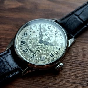 Puede incluir: Reloj de pulsera plateado con una correa de cuero negro. La esfera del reloj presenta n&uacute;meros romanos y un grabado floral intrincado. Tiene una peque&ntilde;a subesfera y una corona texturizada. La correa mide aproximadamente 2 cm de ancho.
