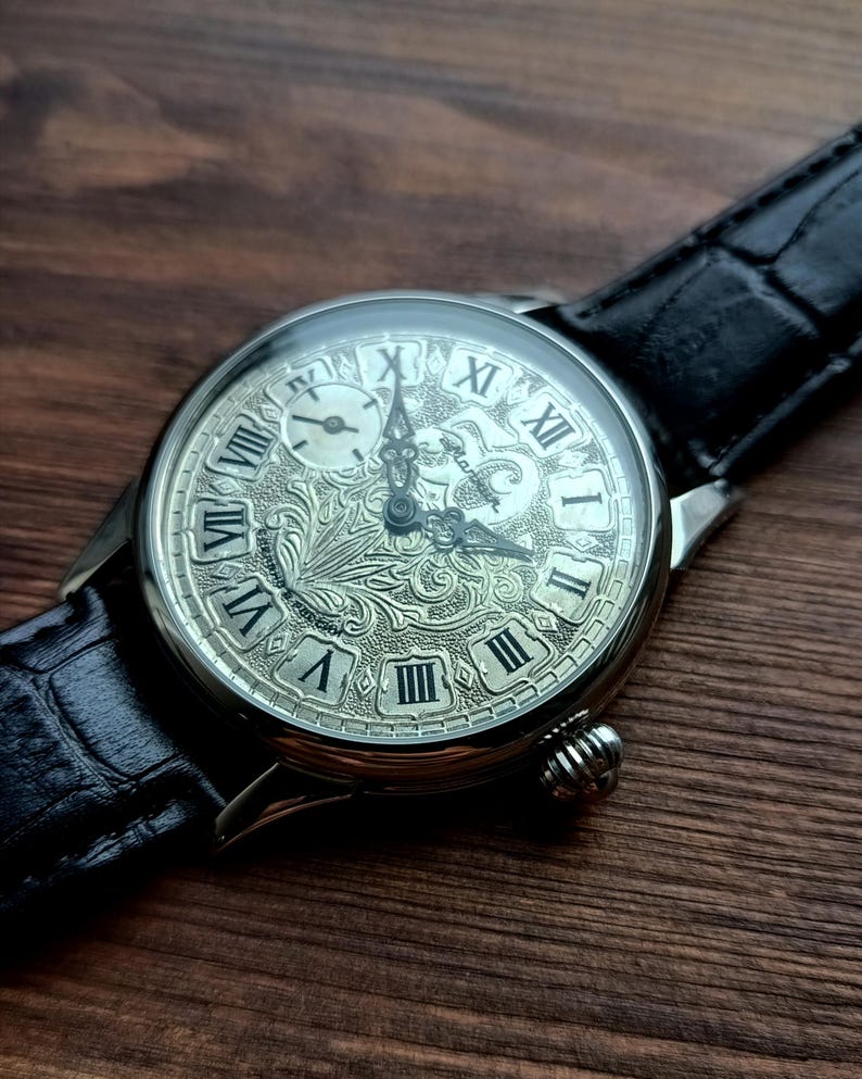 Puede incluir: Reloj de pulsera plateado con una correa de cuero negro. La esfera del reloj presenta n&uacute;meros romanos y un grabado floral intrincado. Tiene una peque&ntilde;a subesfera y una corona texturizada. La correa mide aproximadamente 2 cm de ancho.