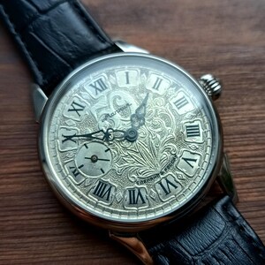 Puede incluir: Un reloj de pulsera plateado con una esfera grabada detallada con n&uacute;meros romanos y una esfera secundaria m&aacute;s peque&ntilde;a. El reloj tiene una correa de cuero negro y un dise&ntilde;o texturizado en la esfera.