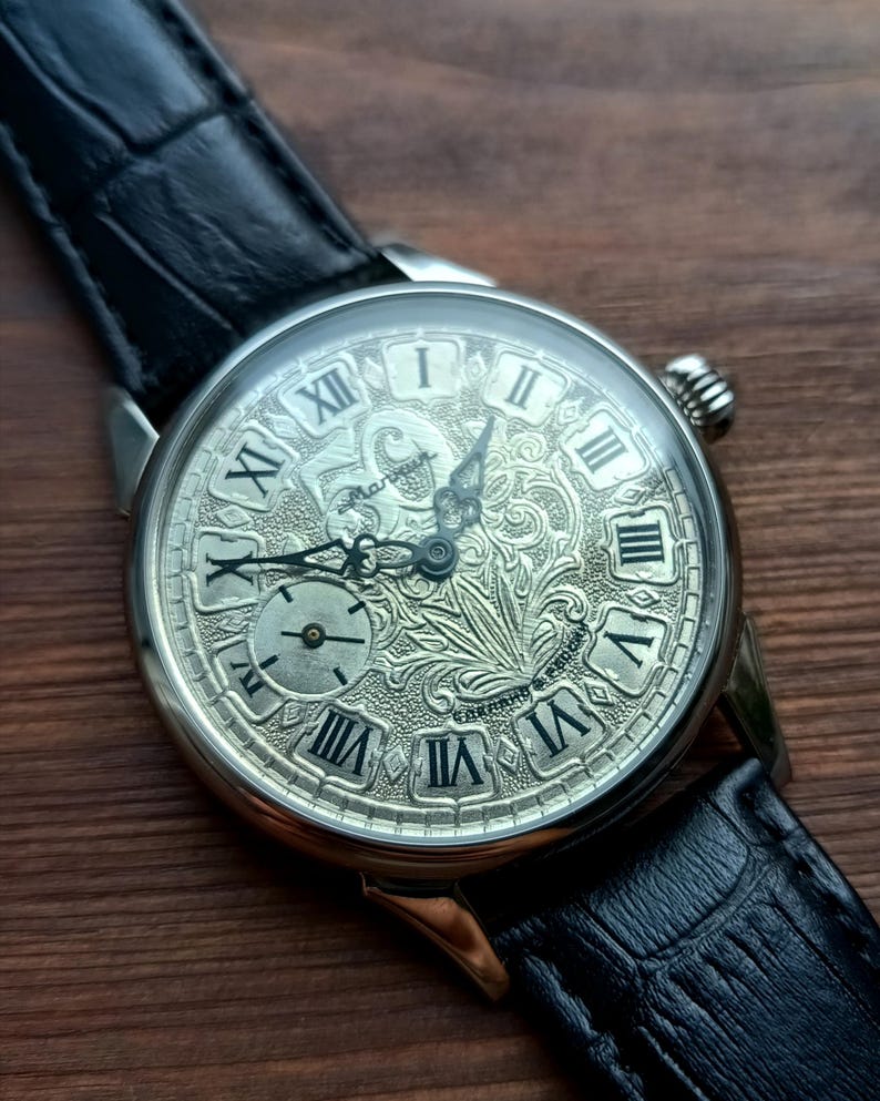 Puede incluir: Un reloj de pulsera plateado con una esfera grabada detallada con n&uacute;meros romanos y una esfera secundaria m&aacute;s peque&ntilde;a. El reloj tiene una correa de cuero negro y un dise&ntilde;o texturizado en la esfera.