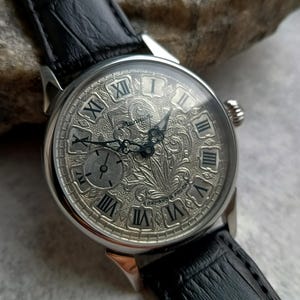Puede incluir: Un reloj de pulsera plateado con una correa de cuero negro. La esfera del reloj presenta n&uacute;meros romanos y un grabado floral intrincado. El reloj tiene una peque&ntilde;a subesfera y una corona texturizada. La correa es de cuero negro.