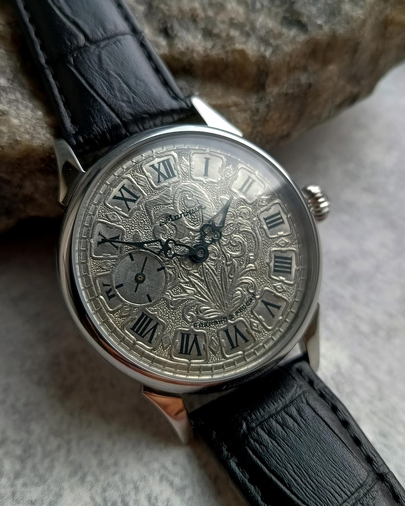 Puede incluir: Un reloj de pulsera plateado con una correa de cuero negro. La esfera del reloj presenta n&uacute;meros romanos y un grabado floral intrincado. El reloj tiene una peque&ntilde;a subesfera y una corona texturizada. La correa es de cuero negro.