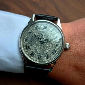 Puede incluir: Reloj de pulsera plateado con una correa de cuero negro. La esfera del reloj presenta n&uacute;meros romanos y un dise&ntilde;o grabado ornamentado. El reloj tiene una peque&ntilde;a subesfera y una corona texturizada. El reloj est&aacute; en una mu&ntilde;eca.