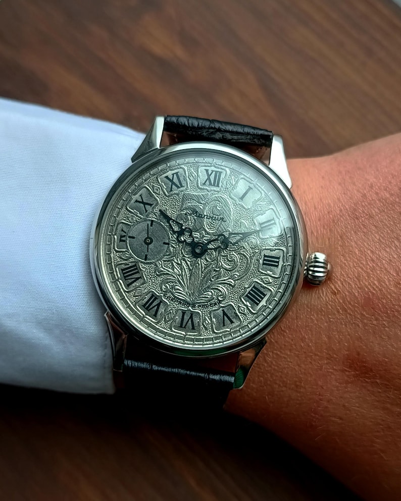 Puede incluir: Reloj de pulsera plateado con una correa de cuero negro. La esfera del reloj presenta n&uacute;meros romanos y un dise&ntilde;o grabado ornamentado. El reloj tiene una peque&ntilde;a subesfera y una corona texturizada. El reloj est&aacute; en una mu&ntilde;eca.