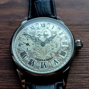 Puede incluir: Reloj de pulsera plateado con una correa de cuero negro. La esfera del reloj presenta n&uacute;meros romanos y un dise&ntilde;o grabado ornamentado. Una esfera secundaria m&aacute;s peque&ntilde;a es visible en el lado izquierdo. El reloj est&aacute; sobre una superficie de madera oscura.