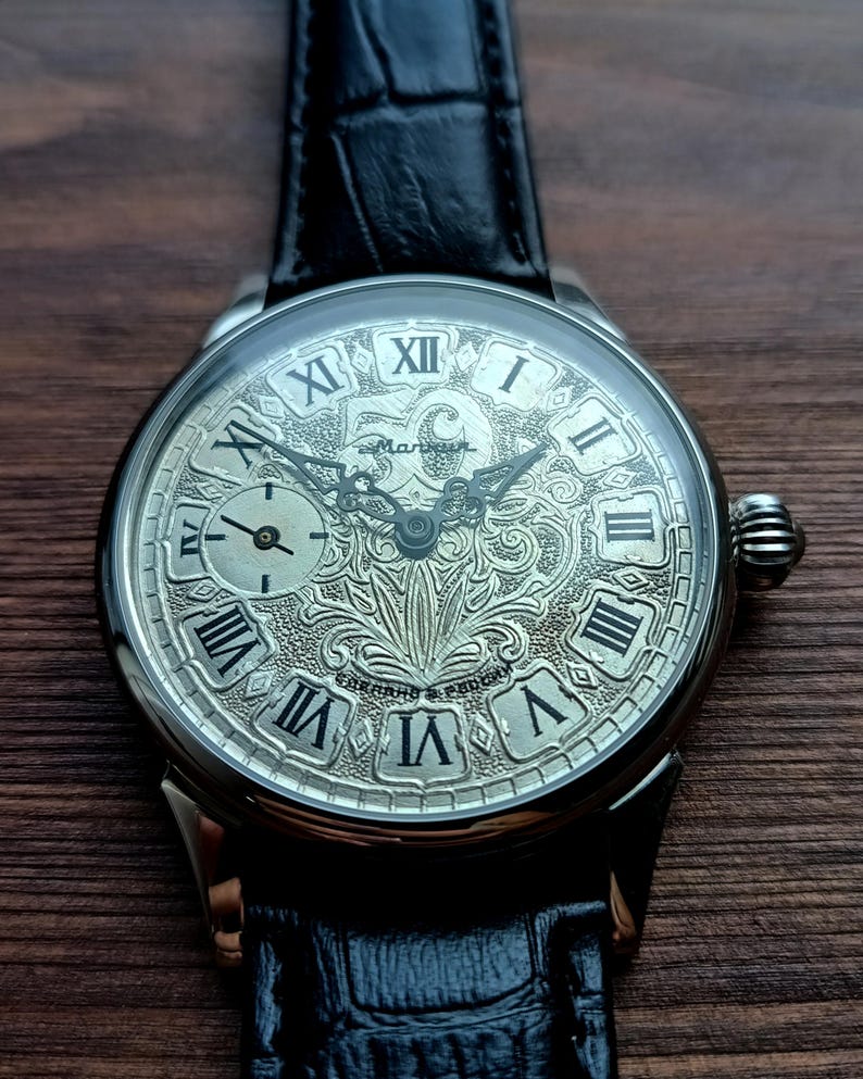 Puede incluir: Reloj de pulsera plateado con una correa de cuero negro. La esfera del reloj presenta n&uacute;meros romanos y un dise&ntilde;o grabado ornamentado. Una esfera secundaria m&aacute;s peque&ntilde;a es visible en el lado izquierdo. El reloj est&aacute; sobre una superficie de madera oscura.