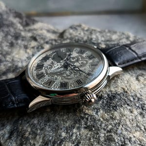 Puede incluir: Reloj de pulsera plateado con una esfera grabada y adornada con n&uacute;meros romanos. El reloj tiene una correa de cuero negro y una corona decorativa. El reloj est&aacute; sobre un fondo gris texturizado. El di&aacute;metro de la esfera es de aproximadamente 4 cm.