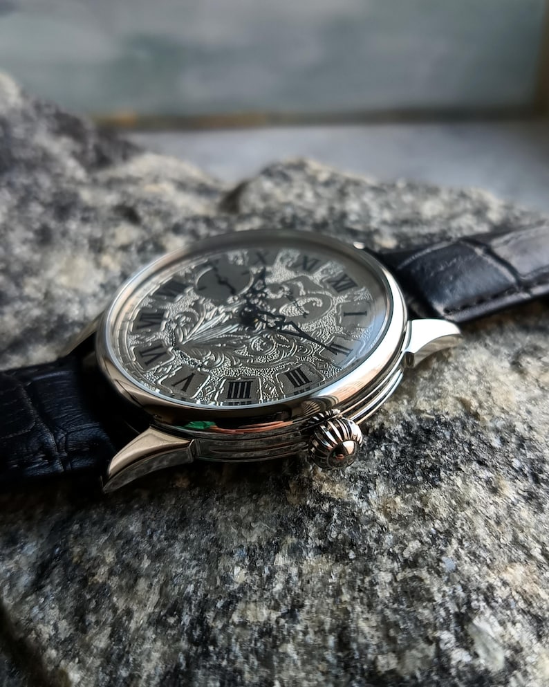 Puede incluir: Reloj de pulsera plateado con una esfera grabada y adornada con n&uacute;meros romanos. El reloj tiene una correa de cuero negro y una corona decorativa. El reloj est&aacute; sobre un fondo gris texturizado. El di&aacute;metro de la esfera es de aproximadamente 4 cm.