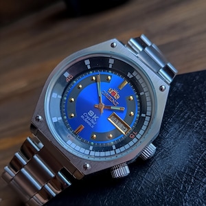 May include: A silver Orient SK Crystal 21 Jewels watch with a blue face and a silver metal band. The watch has a date window and a day of the week window. The watch face has a black outer ring with white markings and a red inner ring with white markings.
