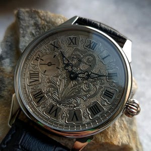 Puede incluir: Reloj de pulsera plateado con correa de cuero negro. La esfera del reloj presenta n&uacute;meros romanos y un grabado floral intrincado. Incluye una peque&ntilde;a subesfera y una corona texturizada.