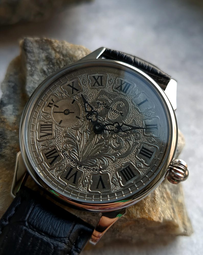 Puede incluir: Reloj de pulsera plateado con correa de cuero negro. La esfera del reloj presenta n&uacute;meros romanos y un grabado floral intrincado. Incluye una peque&ntilde;a subesfera y una corona texturizada.