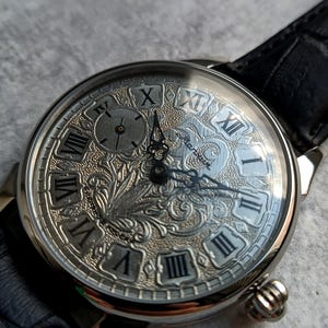 Puede incluir: Reloj de pulsera plateado con una correa de cuero negro. La esfera del reloj presenta n&uacute;meros romanos y un dise&ntilde;o ornamentado y grabado. El reloj tiene una peque&ntilde;a subesfera y una corona texturizada. Las manecillas son negras.