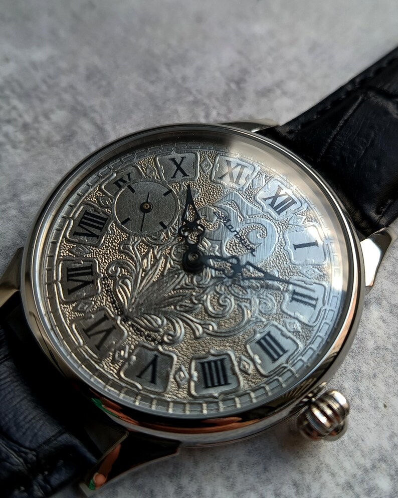 Puede incluir: Reloj de pulsera plateado con una correa de cuero negro. La esfera del reloj presenta n&uacute;meros romanos y un dise&ntilde;o ornamentado y grabado. El reloj tiene una peque&ntilde;a subesfera y una corona texturizada. Las manecillas son negras.