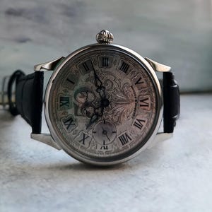 Puede incluir: Reloj de pulsera plateado con correa de cuero negro. La esfera del reloj presenta n&uacute;meros romanos y un dise&ntilde;o grabado. Tiene una peque&ntilde;a subesfera y una corona. Un accesorio cl&aacute;sico y elegante.