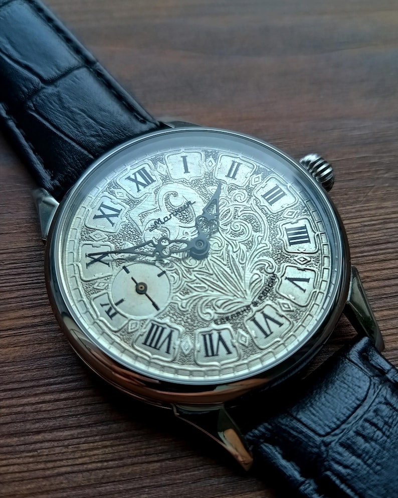 Puede incluir: Reloj de bolsillo plateado con una esfera grabada detallada con n&uacute;meros romanos y una peque&ntilde;a subesfera. El reloj tiene una correa de cuero negro y un dise&ntilde;o ornamentado. Un reloj cl&aacute;sico.